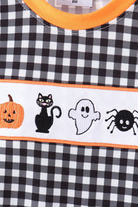 Close-up of a Halloween-themed children's top with a black and white gingham pattern, orange trim, and a white embroidered horizontal band featuring a pumpkin, black cat, ghost, and spider.