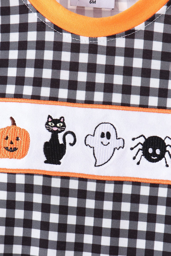Close-up of a Halloween-themed children's top with a black and white gingham pattern, orange trim, and a white embroidered horizontal band featuring a pumpkin, black cat, ghost, and spider.
