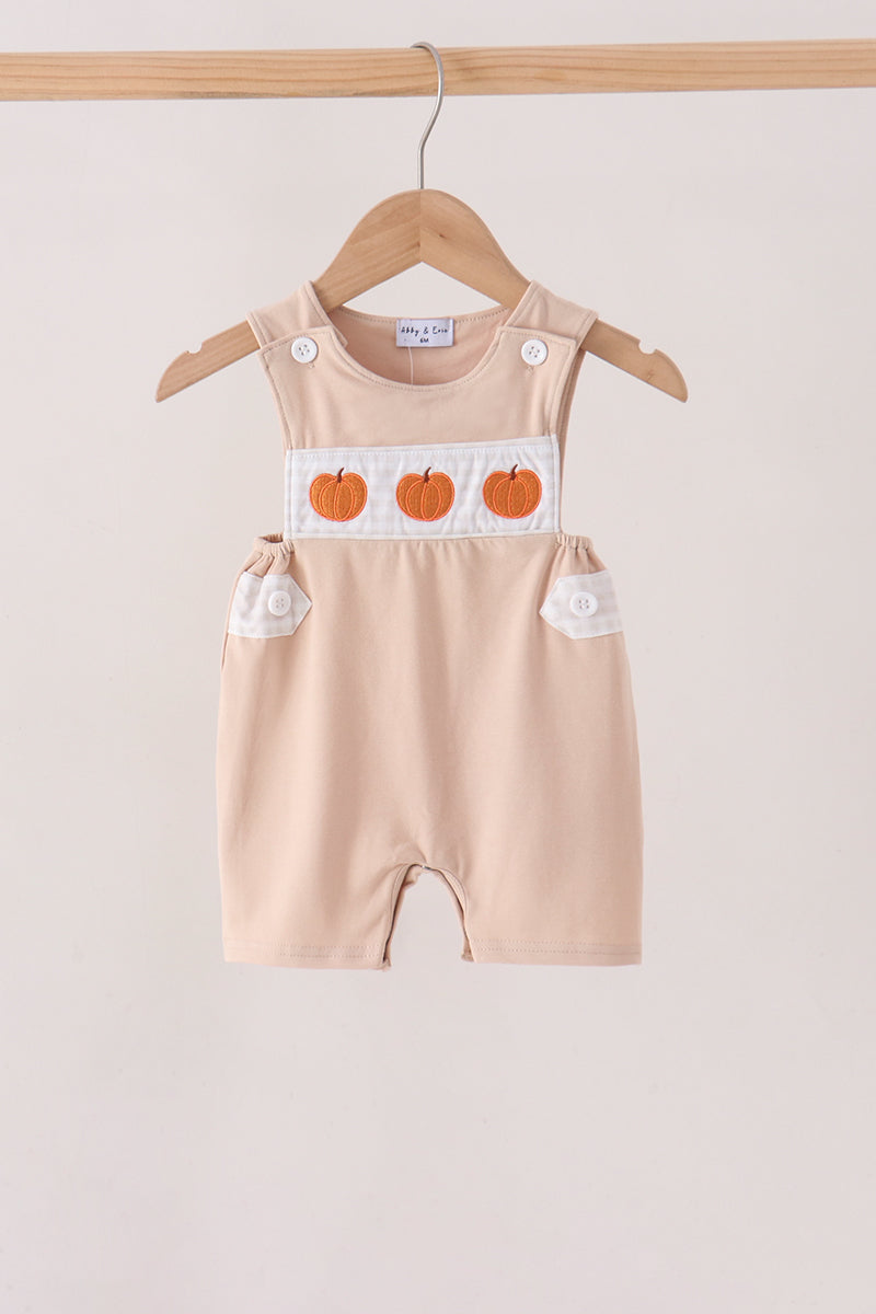 A baby romper in a light peach-beige color, hanging on a wooden hanger. The romper features a white chest panel with three orange embroidered pumpkins and white button details on the shoulder straps and side pockets.