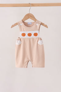 A baby romper in a light peach-beige color, hanging on a wooden hanger. The romper features a white chest panel with three orange embroidered pumpkins and white button details on the shoulder straps and side pockets.