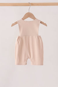 A sleeveless baby romper in a soft blush pink color, hanging on a wooden hanger from a wooden clothing rack against a plain off-white background.
