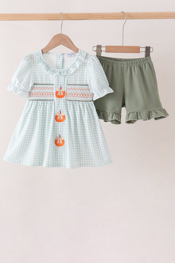 Sage gingham baby dress set with pumpkin embroidery, smocked details, and ruffled solid sage shorts hanging on a wooden rack.