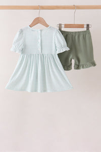 Light sage gingham dress with ruffled sleeves and buttons, paired with sage green ruffle shorts hanging on a wooden rack.