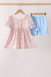 Floral baby dress with pink buttons and blue ruffled shorts set hanging on wooden hangers.