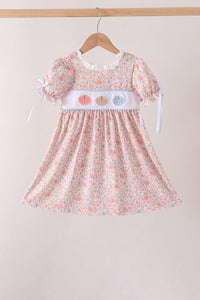 A toddler girl's short-sleeve dress with a pink, white, and blue floral print, featuring a white smocked chest panel with embroidered pumpkins and white ribbon ties, hanging on a wooden hanger against a neutral beige background.