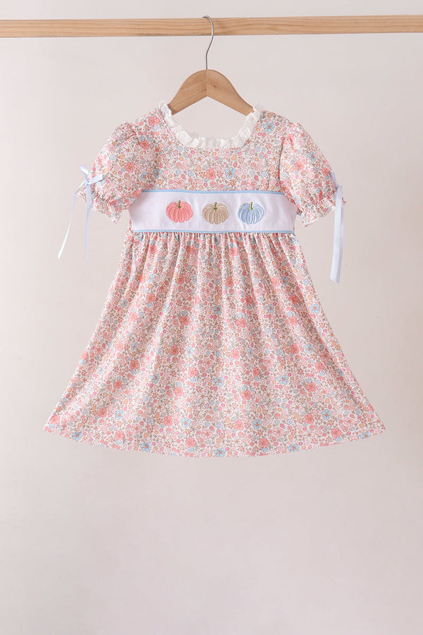 A toddler girl's short-sleeve dress with a pink, white, and blue floral print, featuring a white smocked chest panel with embroidered pumpkins and white ribbon ties, hanging on a wooden hanger against a neutral beige background.
