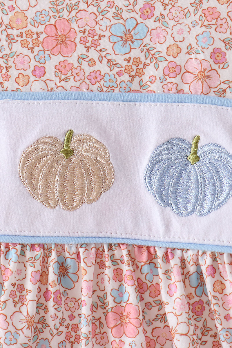 A close-up of a baby dress featuring a white horizontal band with two embroidered pumpkins, one in beige and one in light blue, both with green stems. The dress is decorated with a light blue border and a pink, peach, and blue floral patterned fabric.