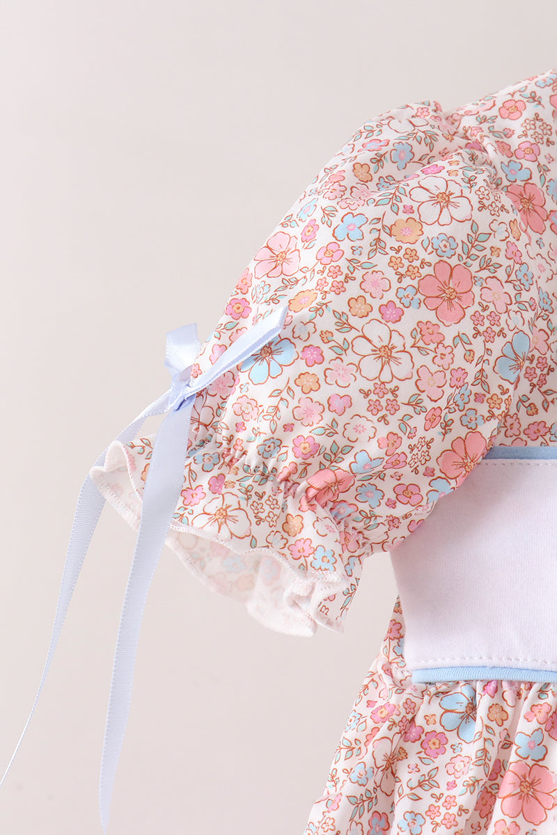 Close-up of a floral-patterned dress with pink, blue, and orange flowers on a cream background, featuring a light blue ribbon bow on the sleeve and a white waistband.