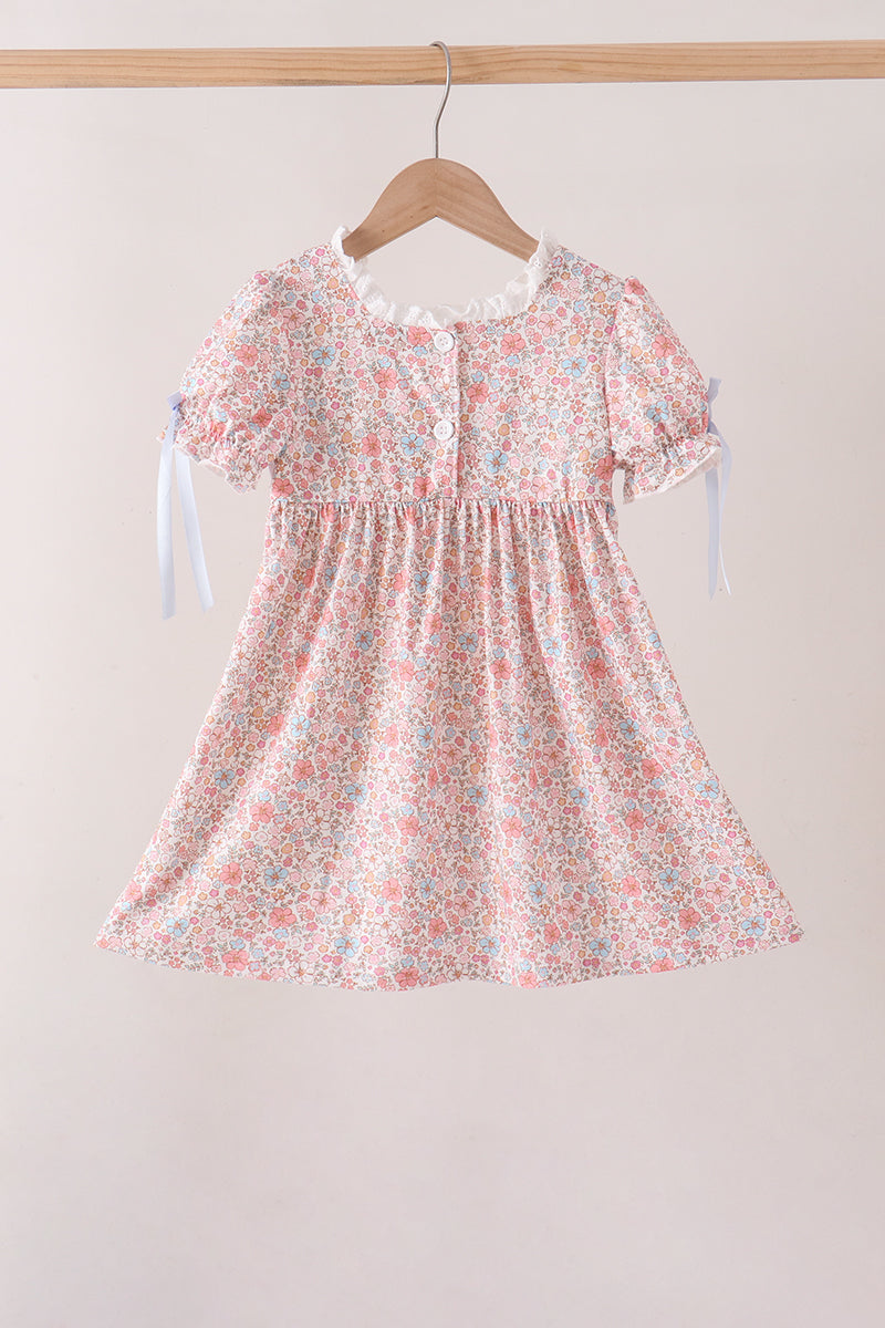 A short-sleeve baby dress with a pink, white, and light blue floral print, hanging on a wooden hanger against a light beige background.
