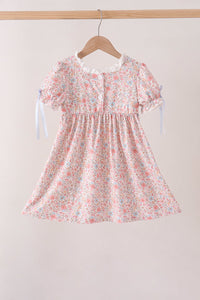 A short-sleeve baby dress with a pink, white, and light blue floral print, hanging on a wooden hanger against a light beige background.