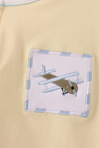 Close-up of a pale yellow fabric garment featuring a square embroidered patch with a vintage airplane design and a light blue striped border.