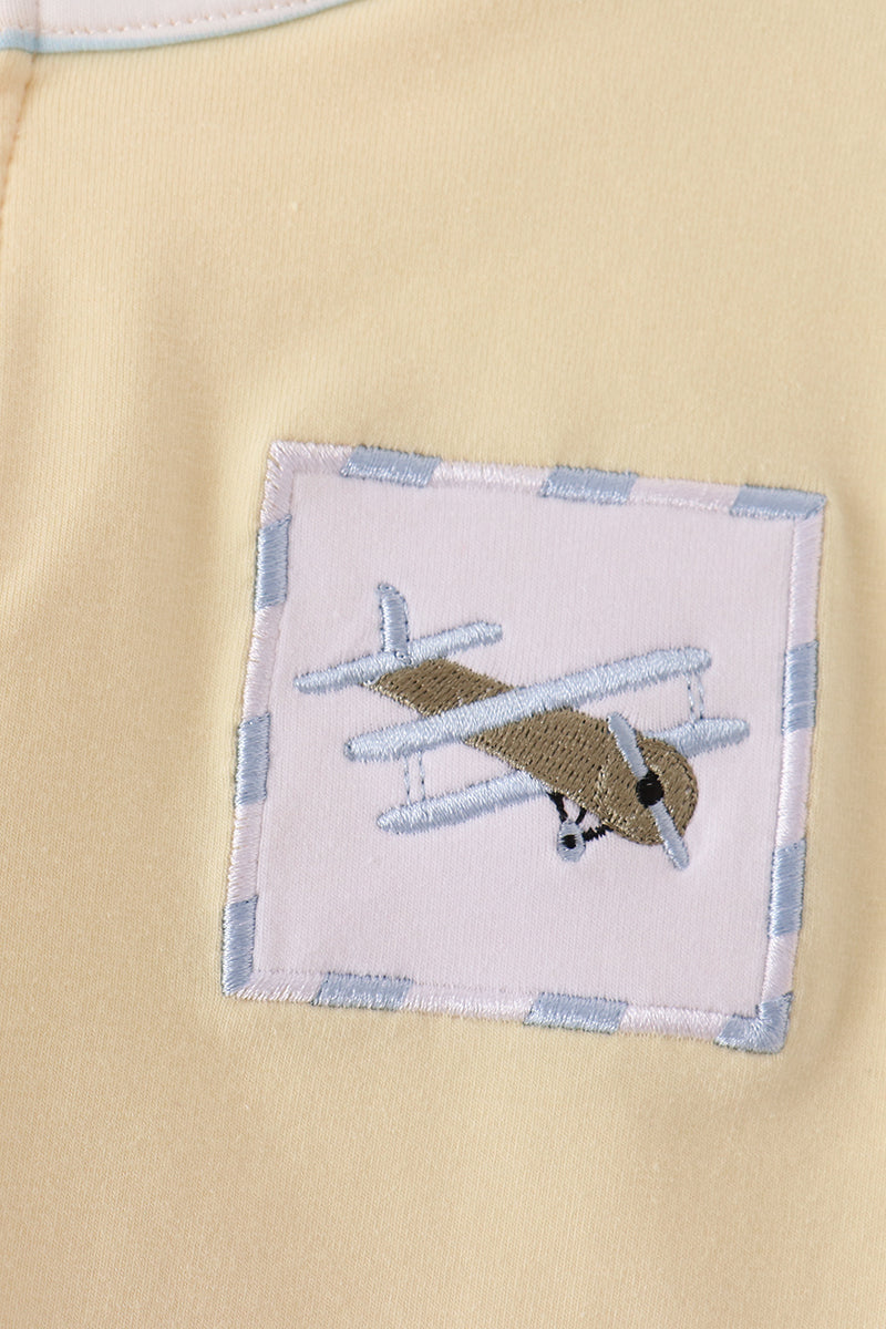 Close-up of a pale yellow fabric garment featuring a square embroidered patch with a vintage airplane design and a light blue striped border.