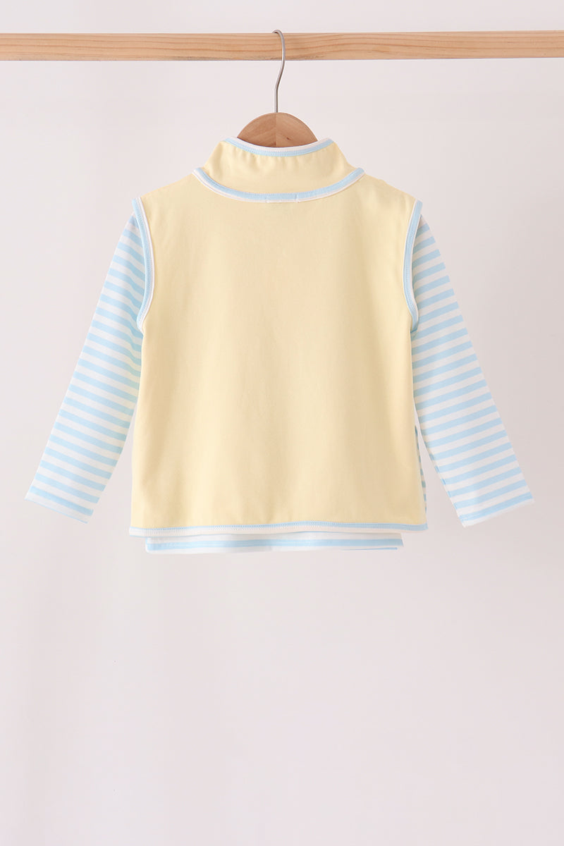 A baby long-sleeve top with a pale yellow mock neck vest body and light blue and white striped sleeves, hanging on a wooden hanger against a light beige background.