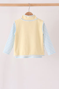 A baby long-sleeve top with a pale yellow mock neck vest body and light blue and white striped sleeves, hanging on a wooden hanger against a light beige background.