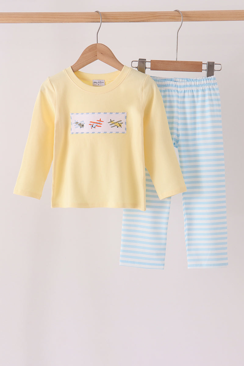 A two-piece children's pajama set hanging on wooden hangers against a light beige background. The set includes a pale yellow long-sleeve top with a small colorful chest graphic and light blue and white striped pants.