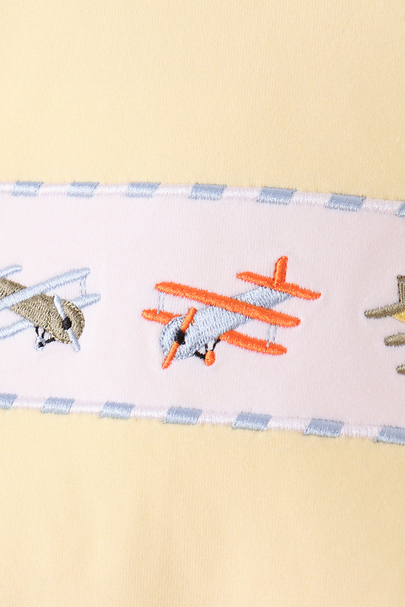 A close-up of a pale yellow fabric item featuring a horizontal embroidered band with cartoon airplanes in orange, gray, and beige on a white background with light blue striped borders.