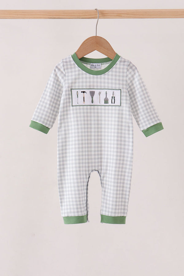 A baby long-sleeve romper in a white and light gray gingham pattern with sage green trim, hanging on a wooden hanger. The romper features a chest graphic of construction tools and is shown against a neutral beige background.
