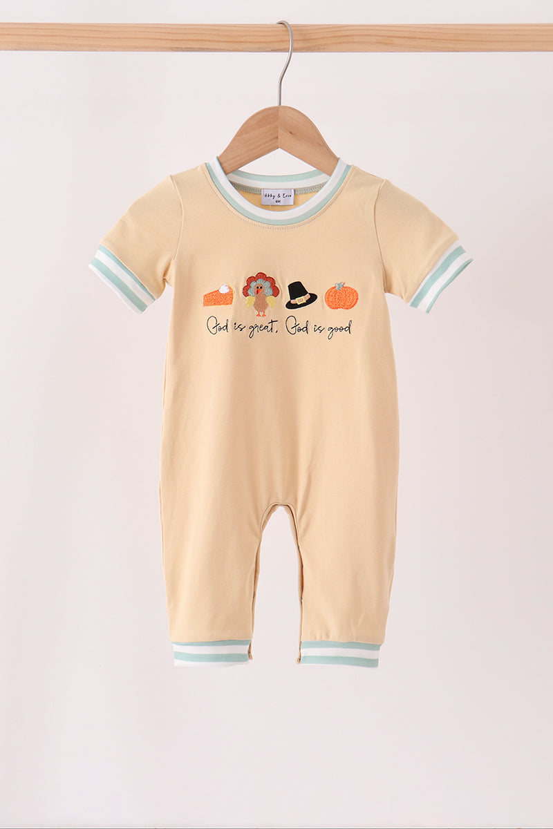 A baby short-sleeve romper in a soft peach-beige color, hanging on a wooden hanger. The romper features light blue and white striped trim on the collar, sleeve cuffs, and leg hems, with a central graphic of autumn-themed icons (pumpkin, witch hat, shoes) and the text 'God be great, God is good'.