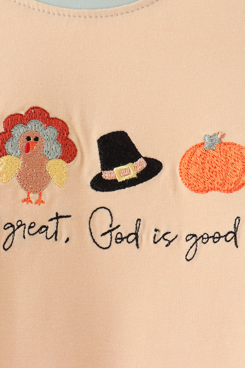 Close-up of a light peach-beige garment with embroidered Thanksgiving motifs, including a turkey, pilgrim hat, pumpkin, and the phrase 'great, God is good' in black script.