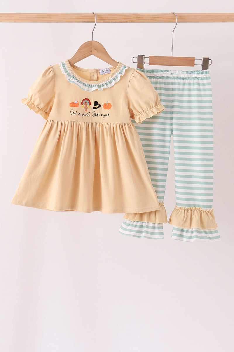 A two-piece baby girl's outfit set hanging on a wooden hanger bar. The set includes a short-sleeve cream-yellow dress with a ruffled collar, pumpkin embroidery, and 'God is good' text, paired with light blue and white striped pants with ruffled hems.