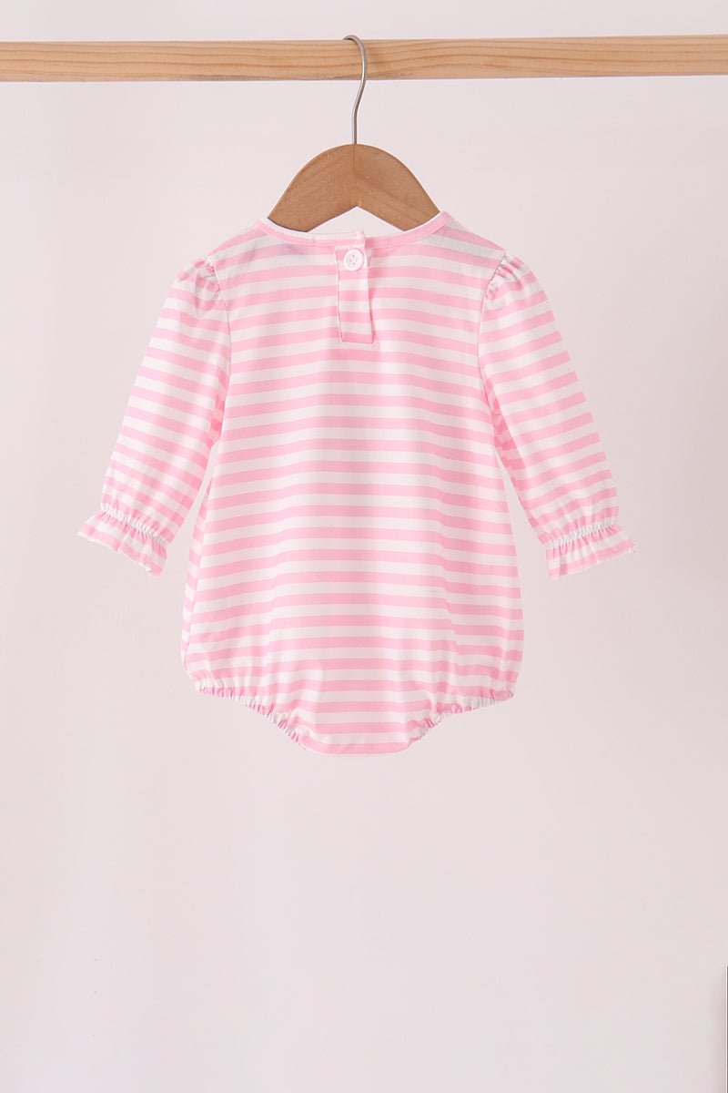 A pink and white striped long-sleeve baby romper hanging on a wooden hanger from a wooden clothes rack against a light pink background.
