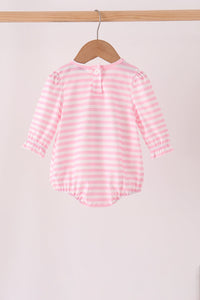 A pink and white striped long-sleeve baby romper hanging on a wooden hanger from a wooden clothes rack against a light pink background.