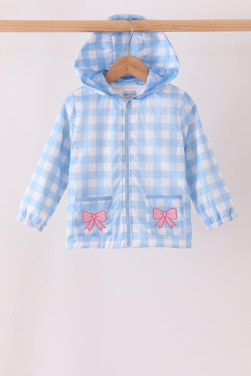 A light blue and white gingham zip-up hooded jacket with pink bow details on the front pockets, hanging on a wooden hanger from a wooden clothes rack against a plain off-white background.