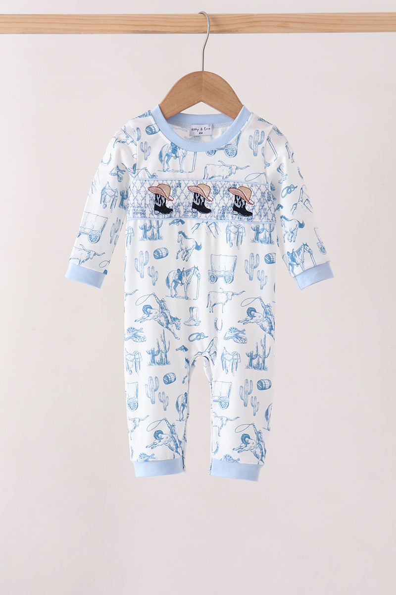 A baby long-sleeve footless romper hanging on a wooden hanger against a plain light beige wall. The romper is white with a light blue cartoon pattern and light blue ribbed cuffs and collar.