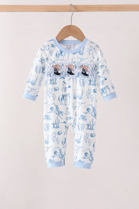 A baby long-sleeve footless romper hanging on a wooden hanger against a plain light beige wall. The romper is white with a light blue cartoon pattern and light blue ribbed cuffs and collar.