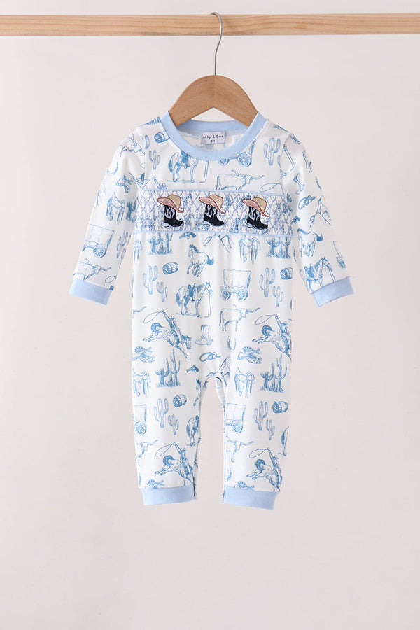 A baby long-sleeve footless romper hanging on a wooden hanger against a plain light beige wall. The romper is white with a light blue cartoon pattern and light blue ribbed cuffs and collar.
