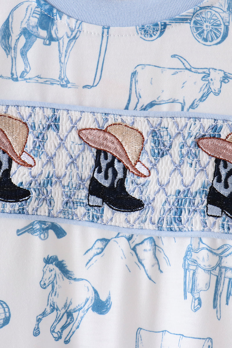 Close-up of a western-themed garment featuring blue line-art illustrations of horses, a steer, and mountains on a white background, with a smocked band displaying embroidered cowboy boots and hats.