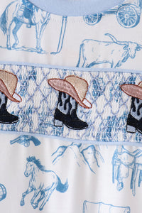 Close-up of a western-themed garment featuring blue line-art illustrations of horses, a steer, and mountains on a white background, with a smocked band displaying embroidered cowboy boots and hats.