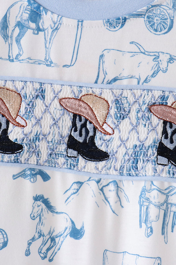 Close-up of a western-themed garment featuring blue line-art illustrations of horses, a steer, and mountains on a white background, with a smocked band displaying embroidered cowboy boots and hats.