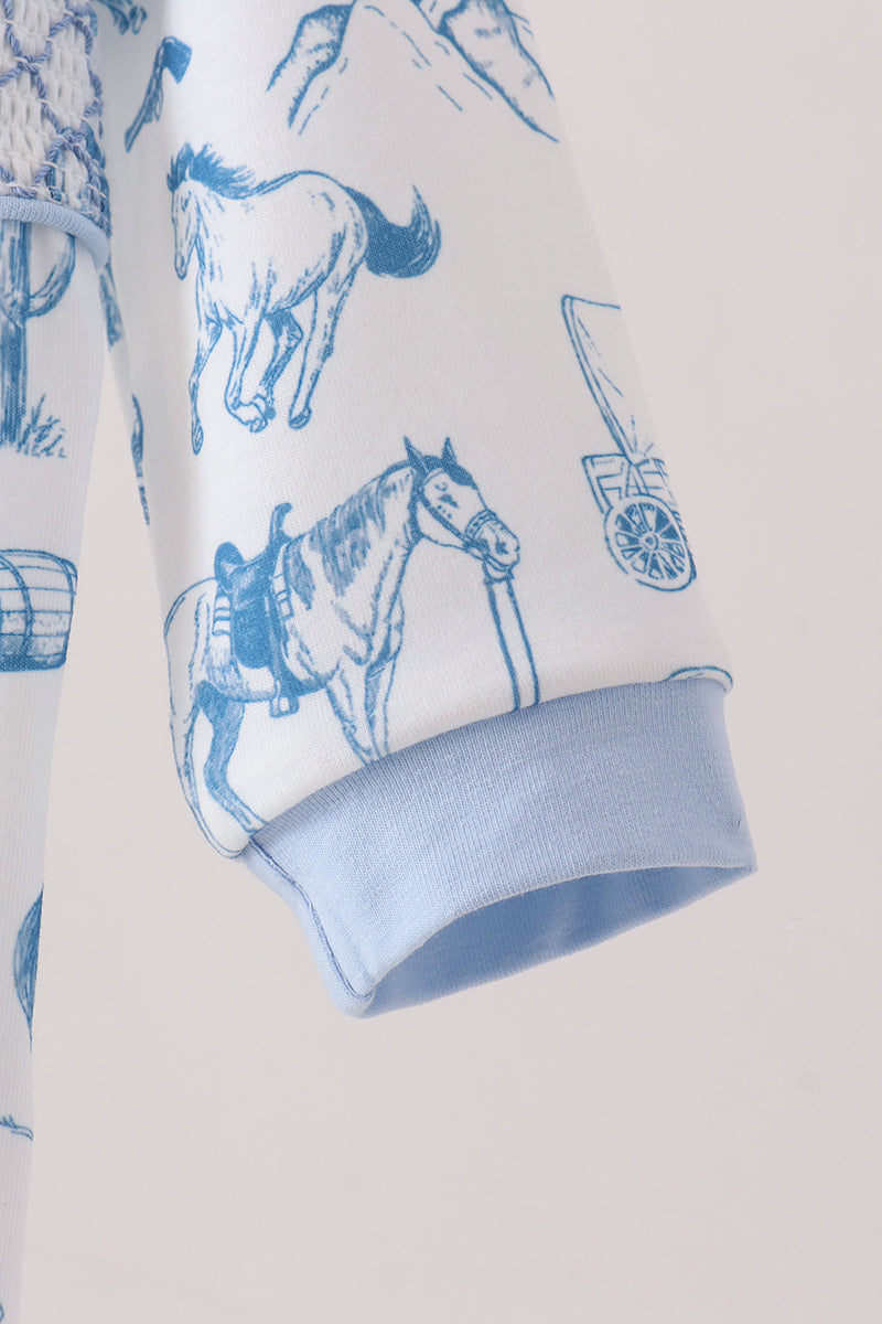 A close-up of a white long-sleeve garment with a blue western-themed print featuring horses, mountains, and a wagon, finished with a light blue ribbed cuff.