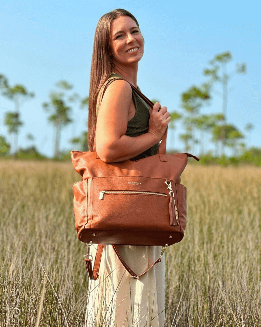 Woman holding cognac diaper bag by top handle in marsh setting, highlighting tote silhouette, gold feet, and adjustable strap