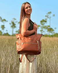 Woman holding cognac diaper bag by top handle in marsh setting, highlighting tote silhouette, gold feet, and adjustable strap