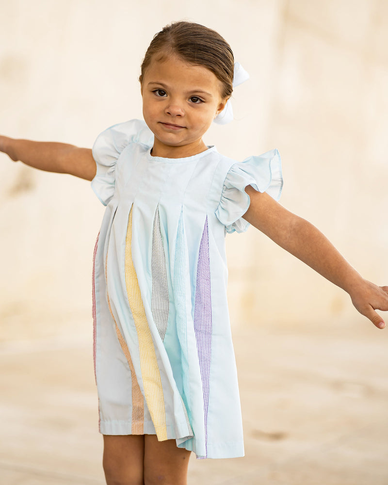 Little girl twirling in a light blue swing dress with flutter sleeves and colorful rainbow pleats down the front.