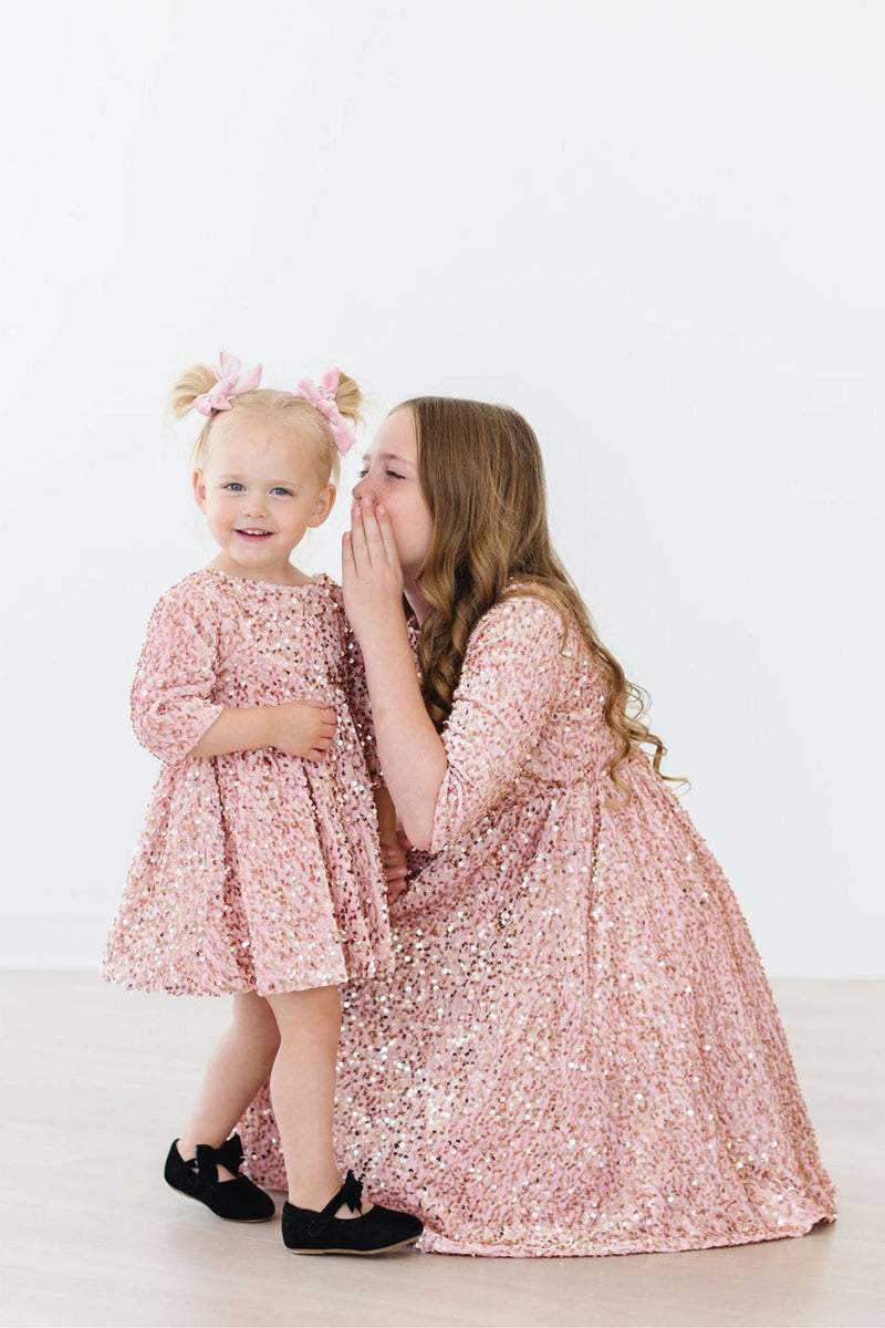 Two girls in the Rose Gold Velvet Sequin Dress, one whispering to the other, showcasing the sparkling design and playful moment.