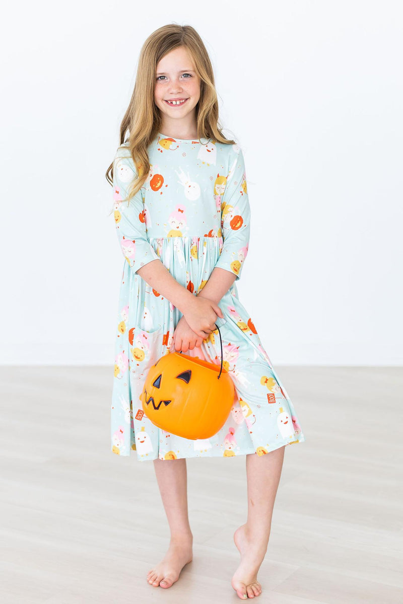A smiling girl in a Ghouls Just Want to Have Fun 3/4 Sleeve Pocket Twirl Dress holding a pumpkin treat bag, ready for Halloween fun and twirling!