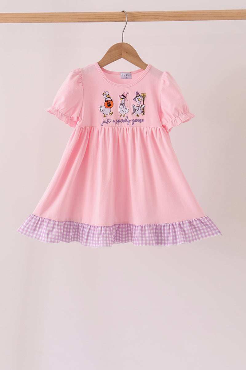 A pink short-sleeve baby dress with a ruffled purple-and-white gingham hem and cartoon unicorn embroidery on the chest, hanging on a wooden hanger against a light beige background.