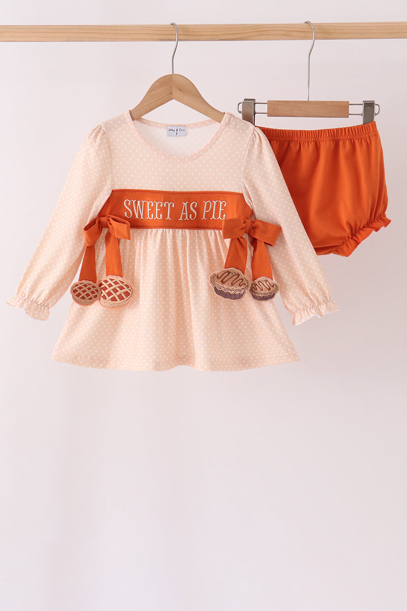 A toddler girl's two-piece outfit set featuring a long-sleeve cream dress with orange polka dots, an orange waistband with 'SWEET AS PIE' text, and orange bow details, paired with matching orange bloomers, hanging on wooden hangers.