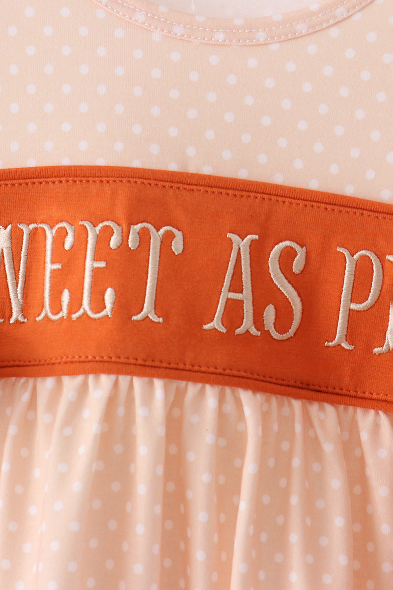 Close-up of a peach and white polka dot dress with a wide orange band across the chest featuring white embroidered text that reads 'SWEET AS P.