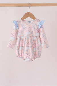 A baby girl's long-sleeve romper with a pink patchwork print featuring light blue ruffle sleeves, hanging on a wooden hanger from a light wood rod against a solid light beige background.