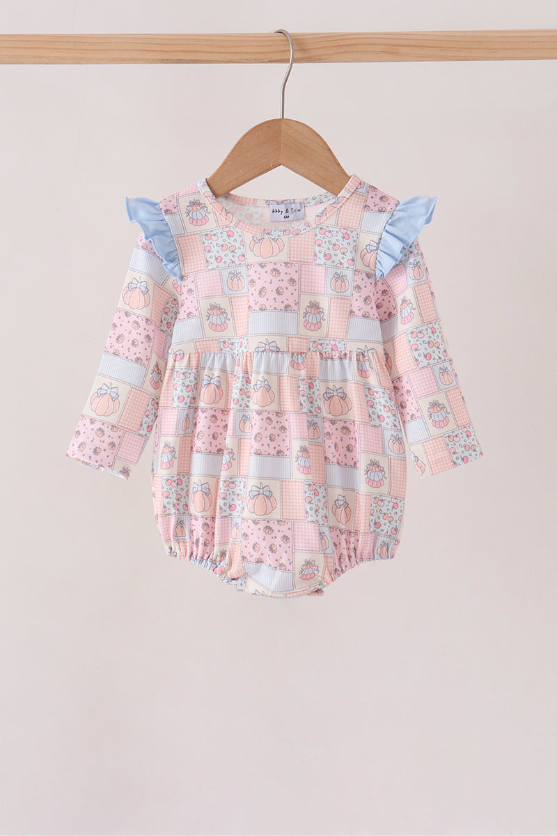 A baby girl's long-sleeve romper with a pink patchwork print featuring light blue ruffle sleeves, hanging on a wooden hanger from a light wood rod against a solid light beige background.