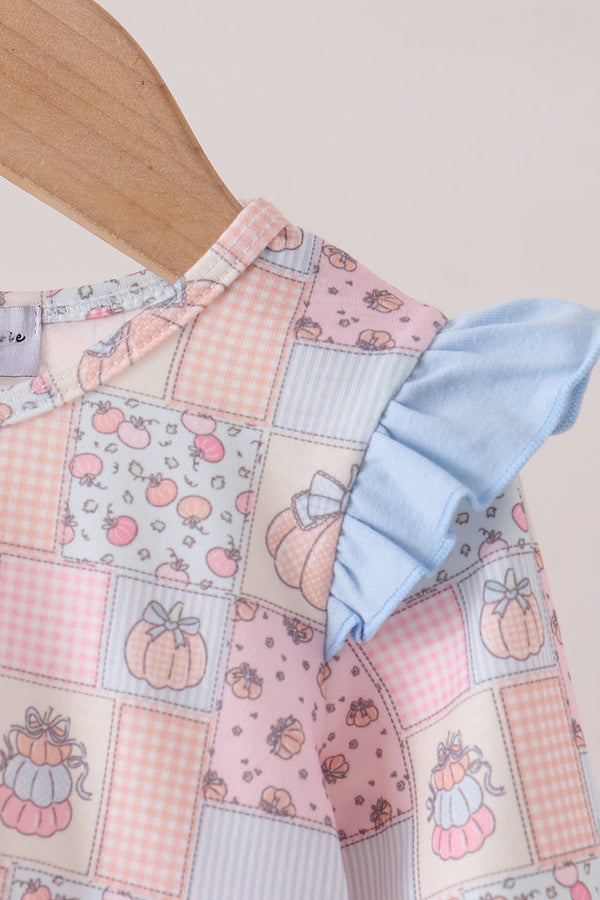 A close-up of a children's dress on a wooden hanger, featuring a pastel patchwork print with gingham, floral, pumpkin, and striped squares, plus a light blue ruffled shoulder detail.