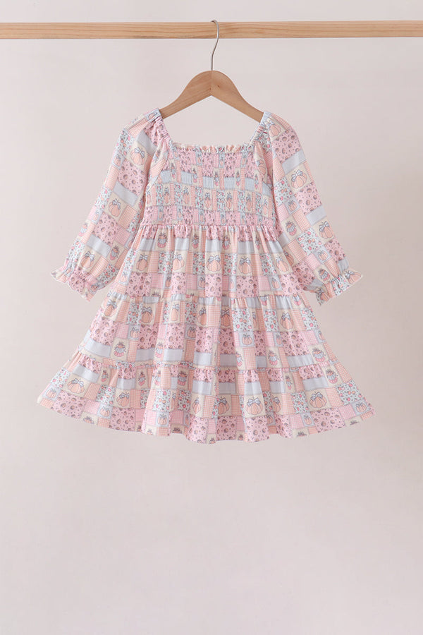 A pink long-sleeve baby dress with a smocked bodice and tiered skirt, featuring a pastel patchwork print with blue, yellow, and white motifs, hanging on a wooden hanger against a light beige wall.