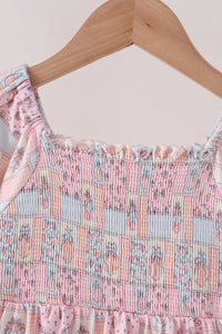 A close-up of a pink and light blue patterned smocked dress top hanging on a wooden hanger against a light beige background.