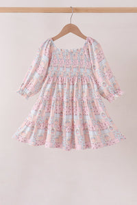 A pink and light blue patterned long-sleeve dress with a smocked bodice, hanging on a wooden hanger against a plain beige background.