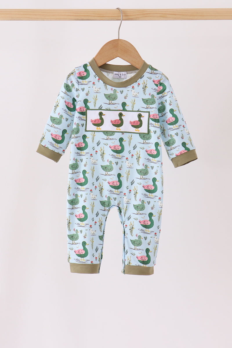 A baby long-sleeve footless romper hanging on a wooden hanger against a light beige background. The romper features a light blue base with an all-over print of green ducks, pink watermelons, and small botanical illustrations, with olive green trim on the collar, cuffs, and hems.