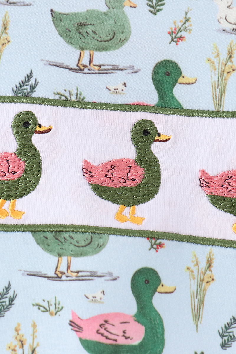 A close-up of a fabric or wallpaper pattern featuring embroidered-style ducks in green and pink on a pale blue background, with a white horizontal band across the center.